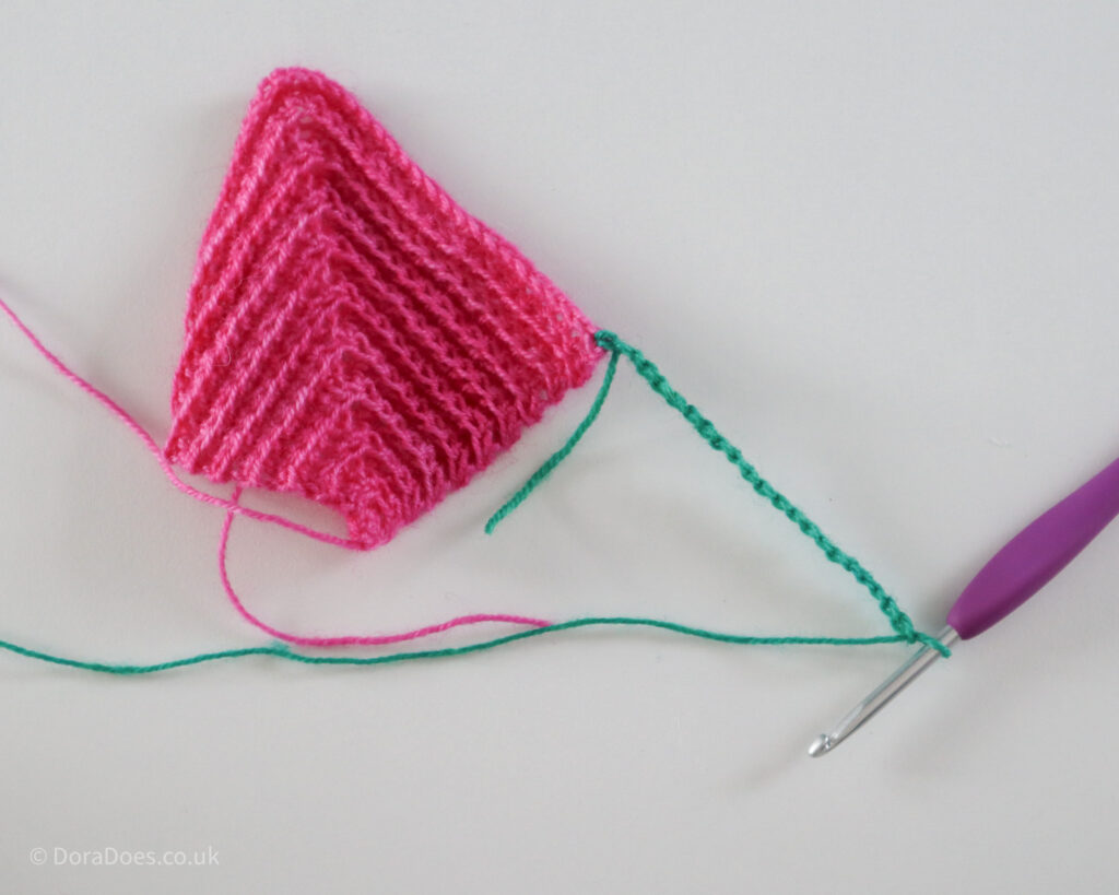 A chain of stitches in green yarn is attached to a pink mitred square showing the beginning of the joining process.