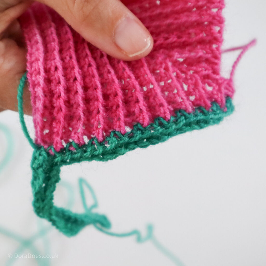 A close up of a row of green crochet stitches worked into the row ends of a pink mitred square.