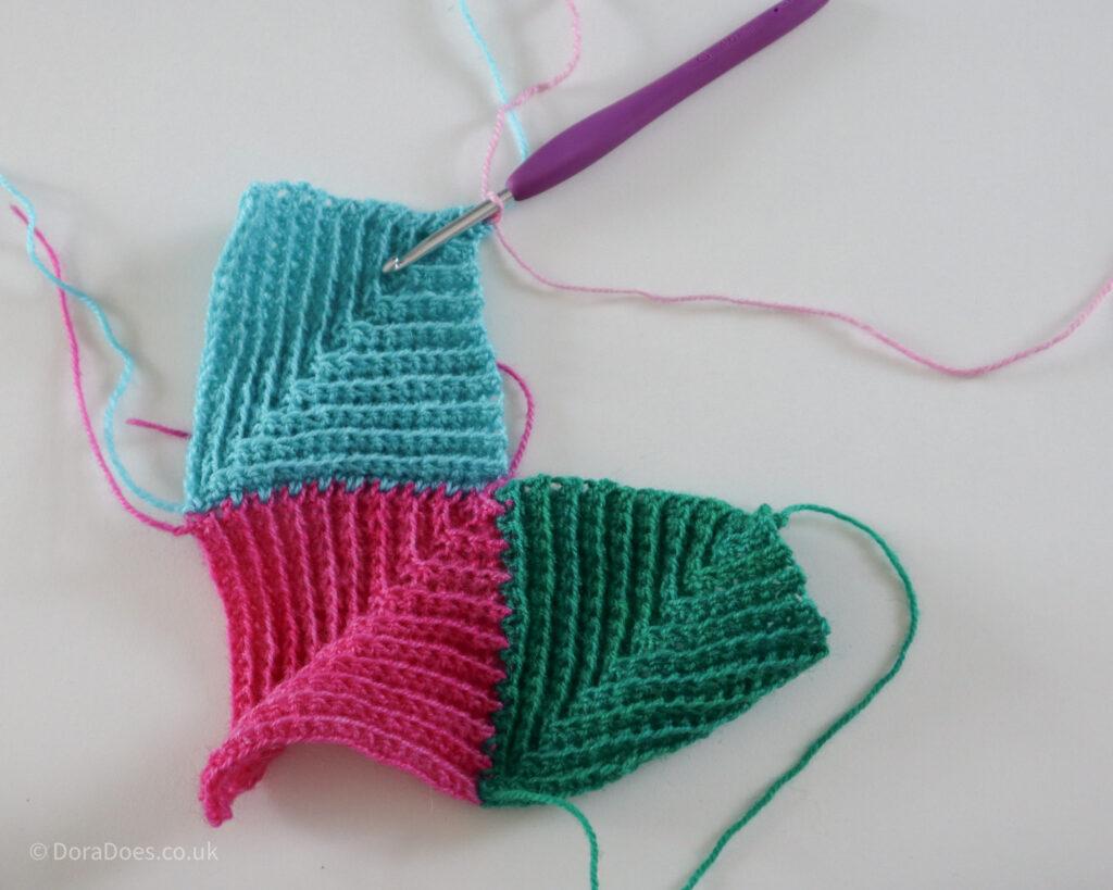 Pink yarn had been joined to the corner of a turquoise crochet mitred square showing the first step of joining a fourth mitred square to a group of three.