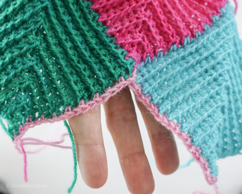 A close up of the first row of a final, pink crochet mitred square, the row worked into the row ends of two previously joined crochet squares.