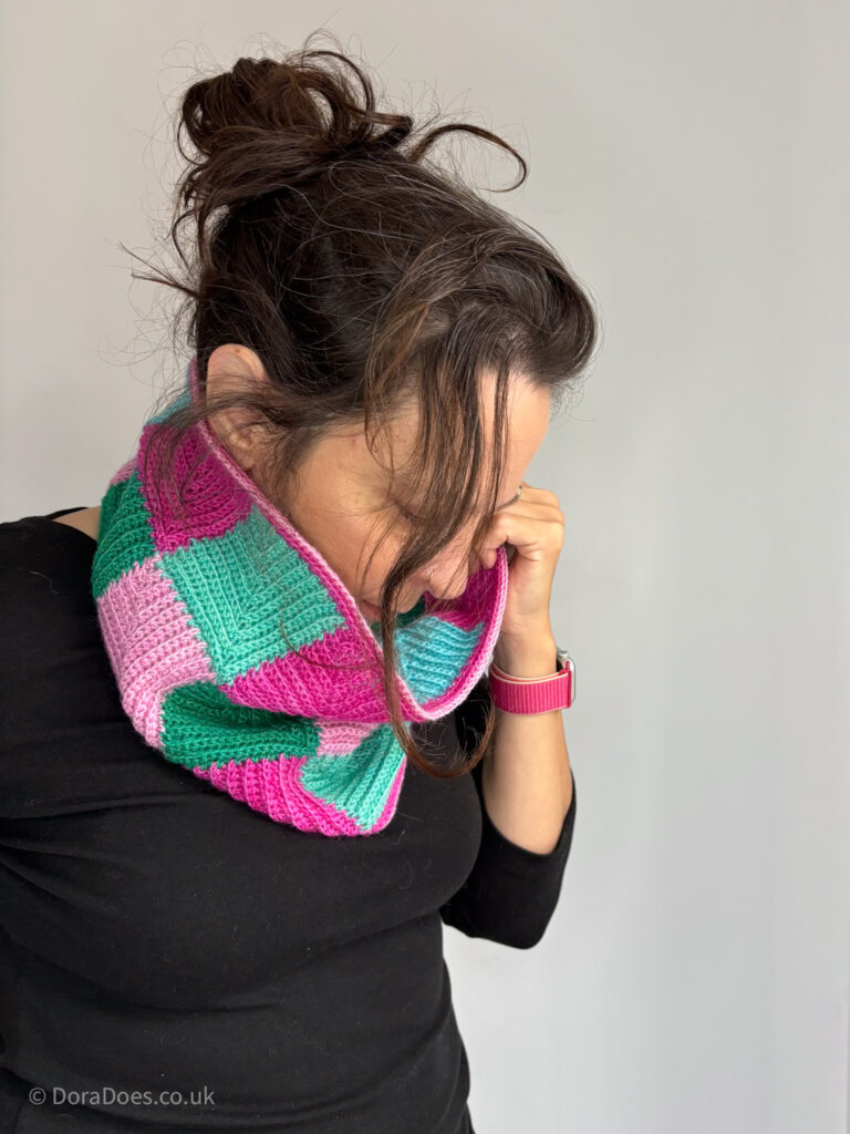 Dora wears a crochet cowl made from mitred squares in pinks and teals.
