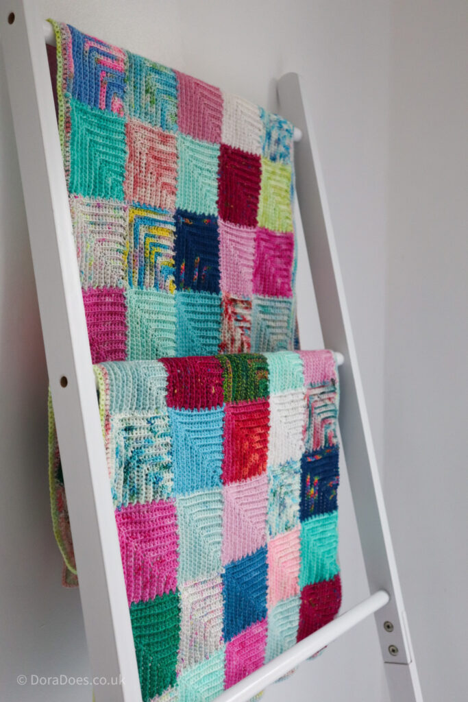 A patchwork mitred square crochet wrap hangs over a blanket ladder.