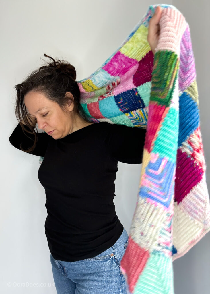 Dora in process of wrapping up on a large crochet wrap made from mitred squares in a multitude of colours. One end of the wrap is held towards the screen and looks disproportionately large.