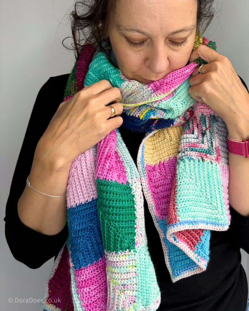 Dora has a large crochet wrap made from mitred squares wrapped around her neck and is looking cosy.