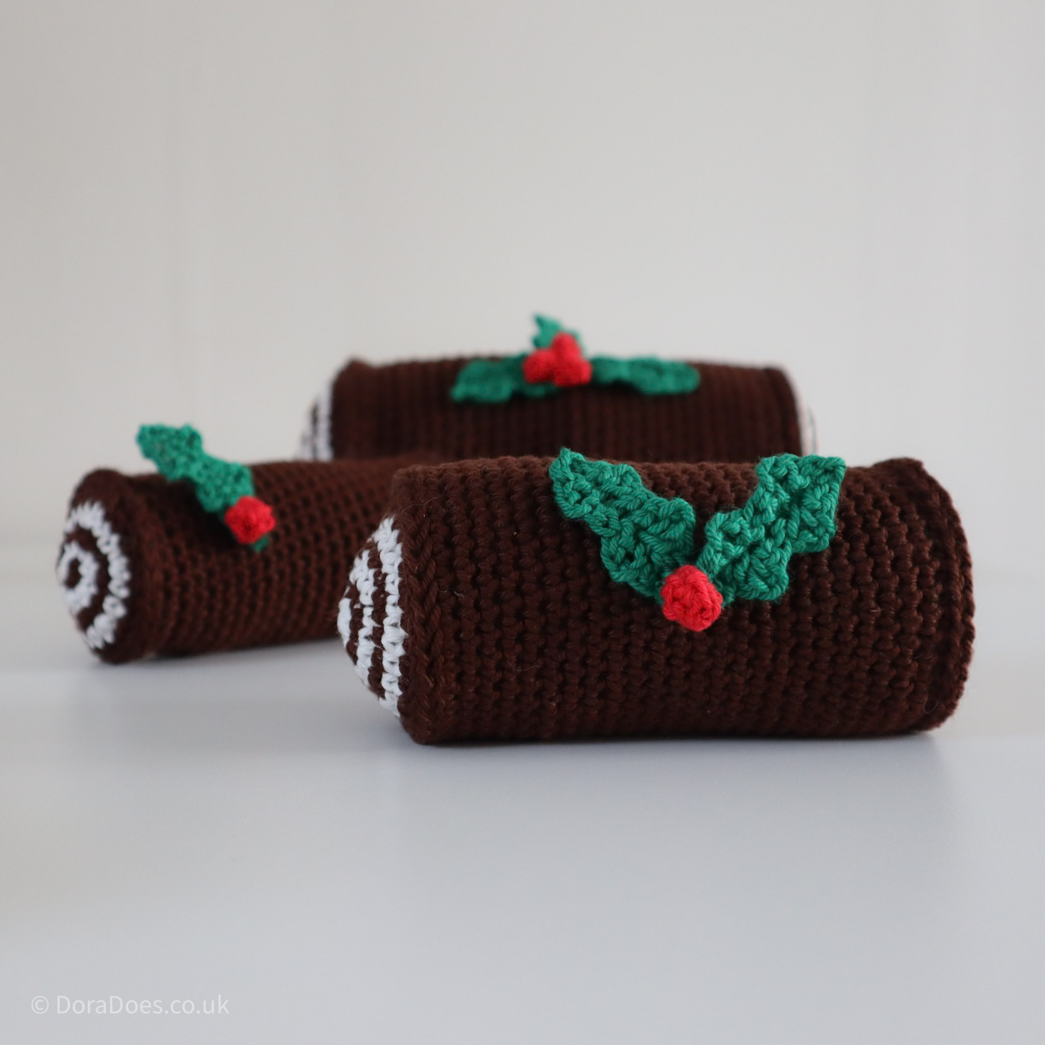 Three crochet Yule logs of different sizes. The medium size decorative crochet log is in focus in the foreground and has two crochet holly leaves, joined with a crochet berry, decorating it.
