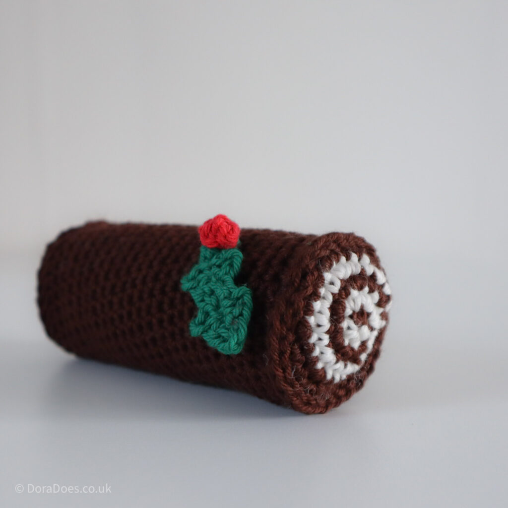 A small crochet Yule log with a single holly leaf and berry decoration seen close up at an angle.