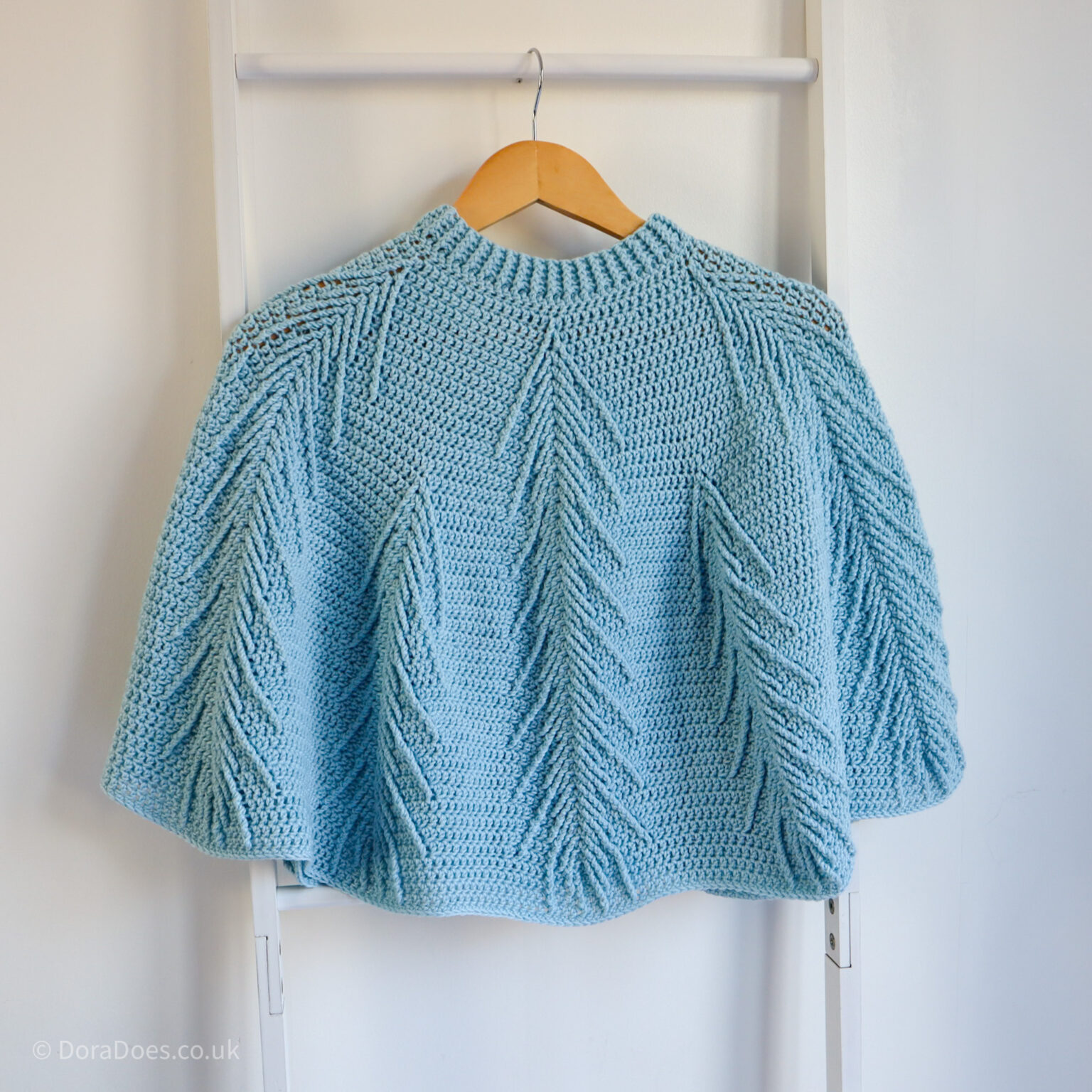 The Falling pines crochet poncho seen from the front hanging on a blanket ladder against a white wall.