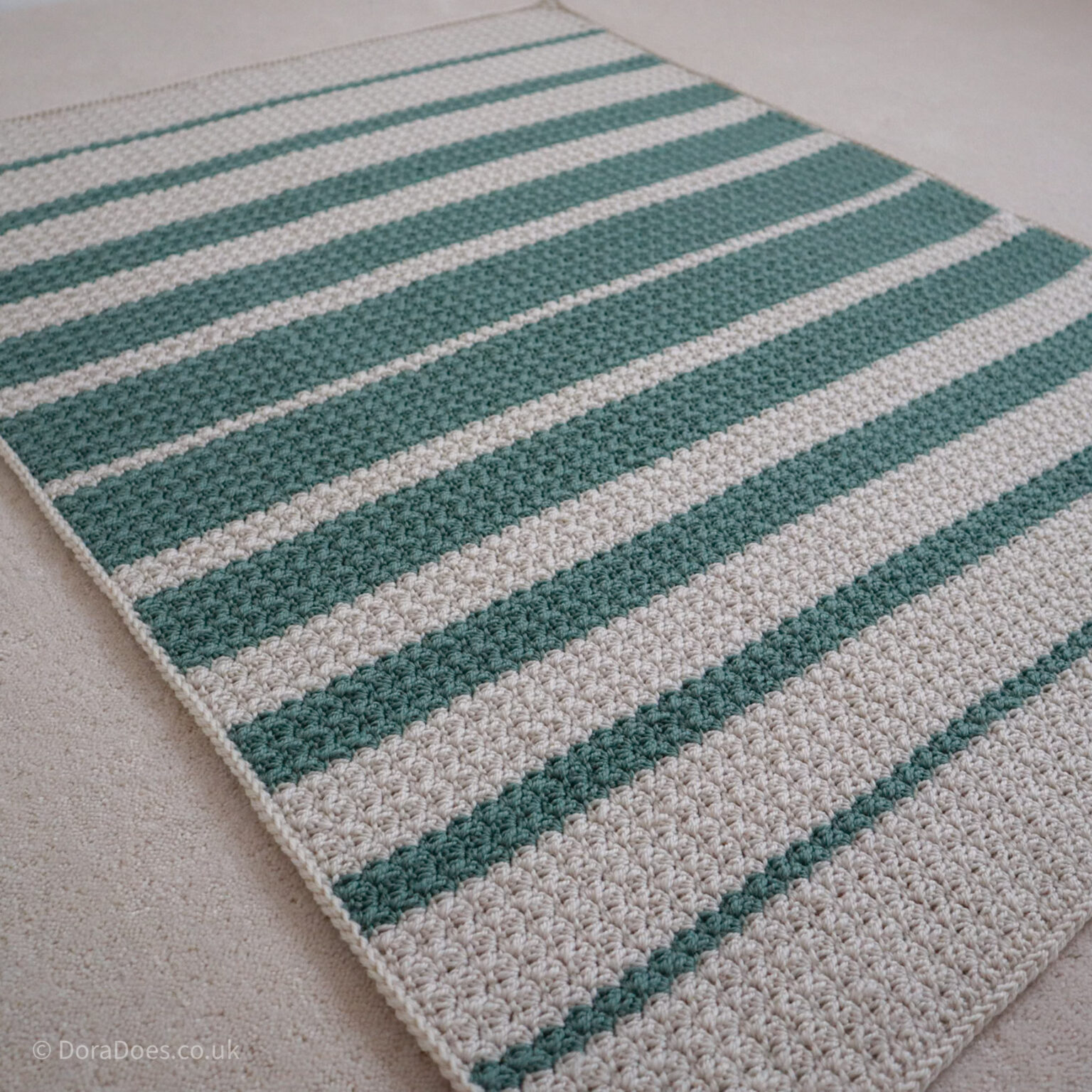 An angled view from above of a textured crochet blanket made with asymmetrical stripes in sage green and natural cream wool. Seen at an angle laying on a cream carpet.