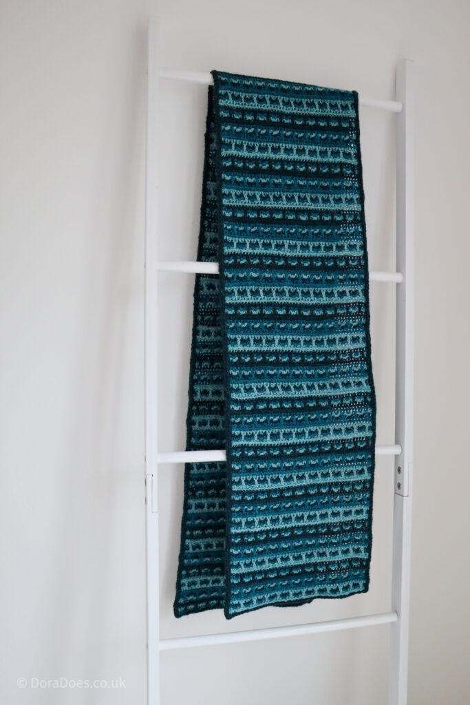 A large crochet wrap in three shades of teal is draped over a blanket latter, showing it’s unusual check pattern.