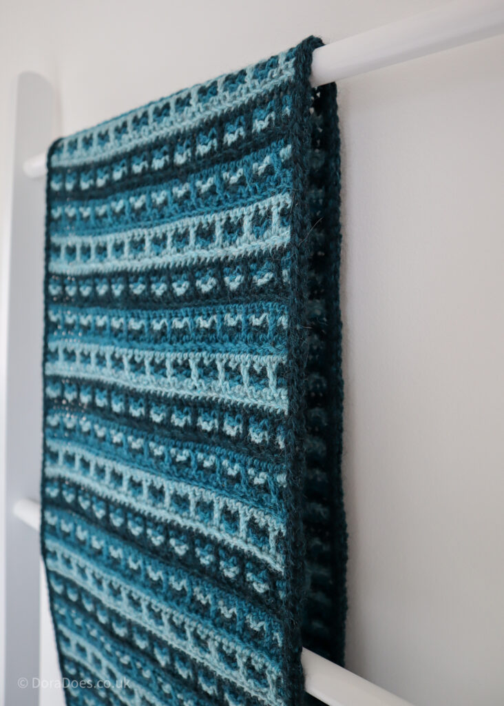 A close up of the top of a crochet scarf as it is draped over a blanket ladder. The wrap has a checked woven effect stitch pattern made in three shades of teal yarnadelic sport weight yarn from John Arbon Textiles.