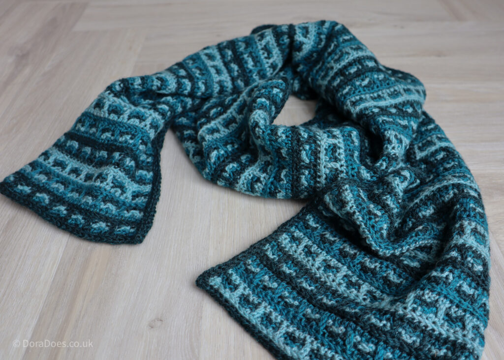 A crochet scarf in three colours of teal lays coiled on the floor. The crochet stitch pattern makes a checked tweed effect.