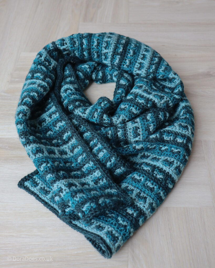 A crochet scarf in three shades of teal is coiled up double on the floor, looking cozy and inviting.