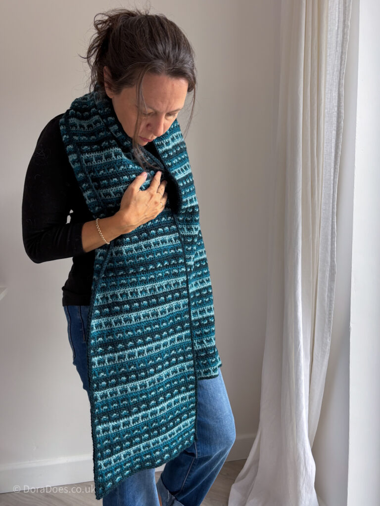 A woman wears a long crochet scarf draped over her shoulders The scarf is a woven plaid pattern with three shades of teal.