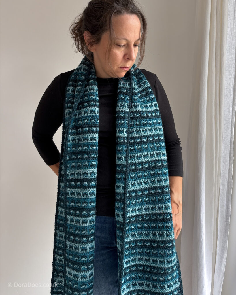 Dora wears an oversized crochet scarf draped around her neck. IT is a tweed style checked plaid pattern made with three shades of teal.