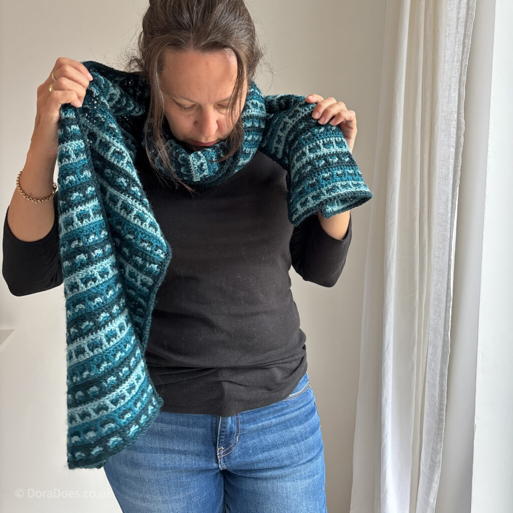 Dora has a crochet scarf with a checked pattern wrapped around her neck, paired with a black top and jeans.
