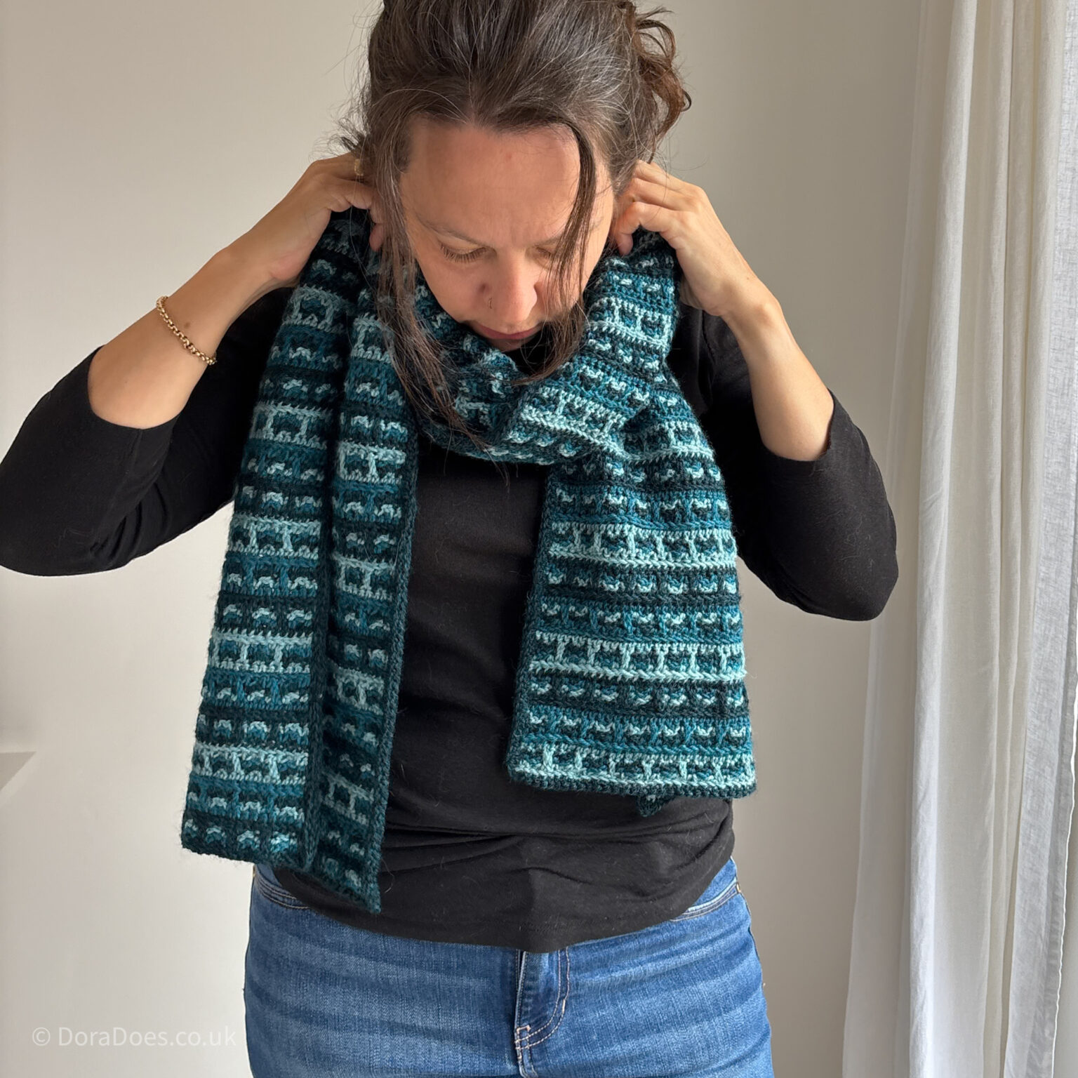 Dora dusts the crochet scarf, made from tonal teals as it curls around her neck, looking cozy.