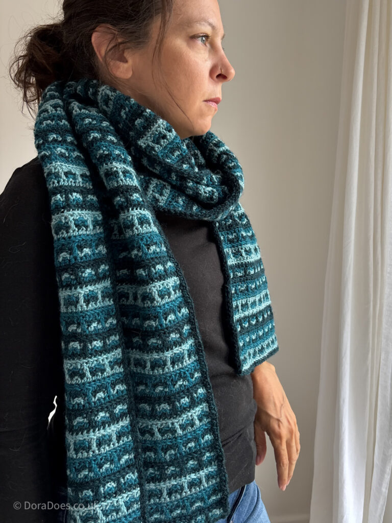 Dora wears a crochet wrap made from three shades of blue green in a checked interlocking crochet stitch pattern.