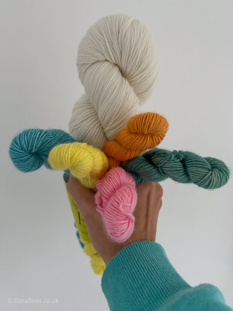 A hand holds 6 skeins of yarn. One large undyed skein of merino wool and 5 mini skeins in pink, green, yellow, blue and orange.