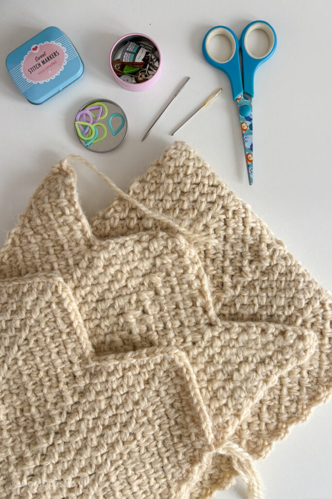 A close up of a zig zag crochet scarf laid flat with the two ends together ready to be joined. A pair of scissors, yarn needles and various stitch makers in small tins are placed beside the scarf.