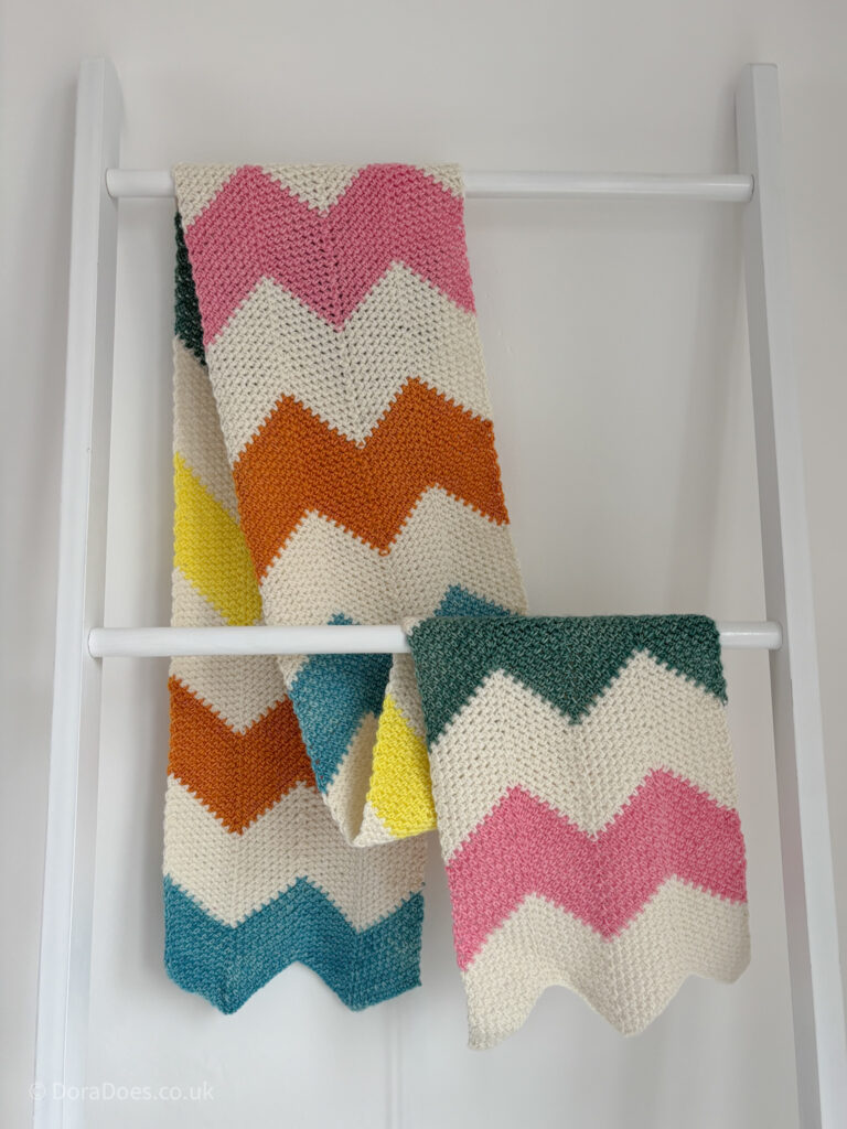 A stripy zig zag scarf is draped over a blanket ladder. The scarf alternates wide stripes of cream merino wool with brightly coloured shades in plant typed yarn.
