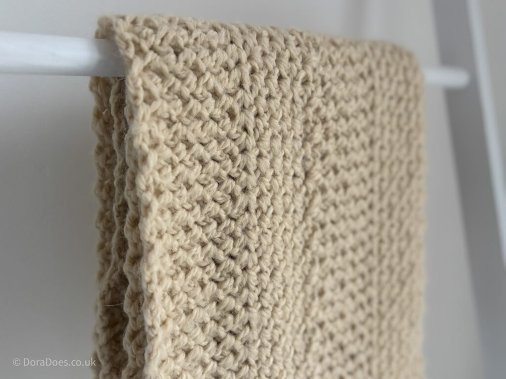 A cream crochet infinity scarf is draped over a rail. The chevron pattern is somewhat disguised by the single colour of yarn used. It looks like a fisherman knit look stitch pattern.