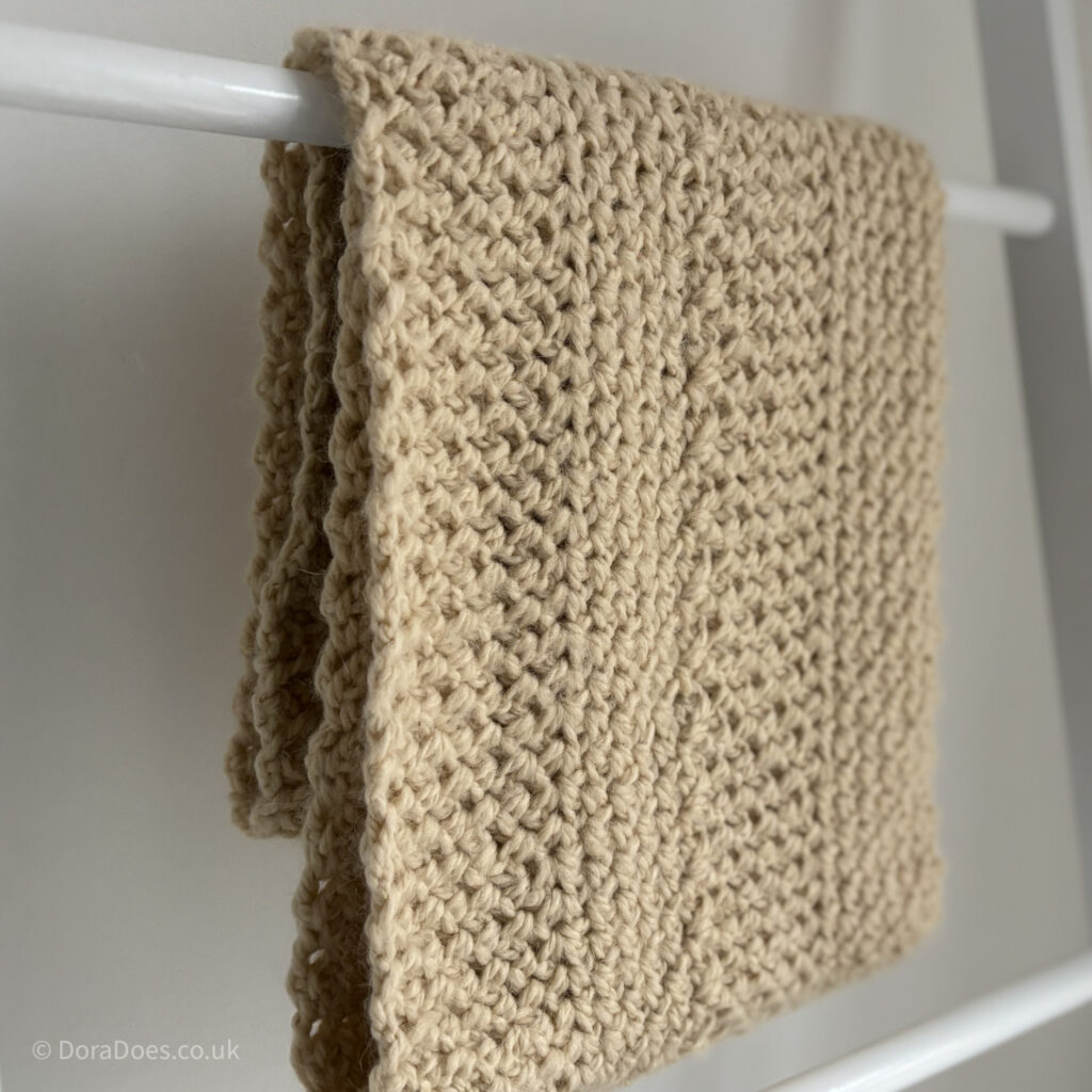 A natural ecru coloured crochet scarf is gilded and draped over a blanket ladder. The zig zag texture of the crochet stitches gives a traditional Aran crochet look.