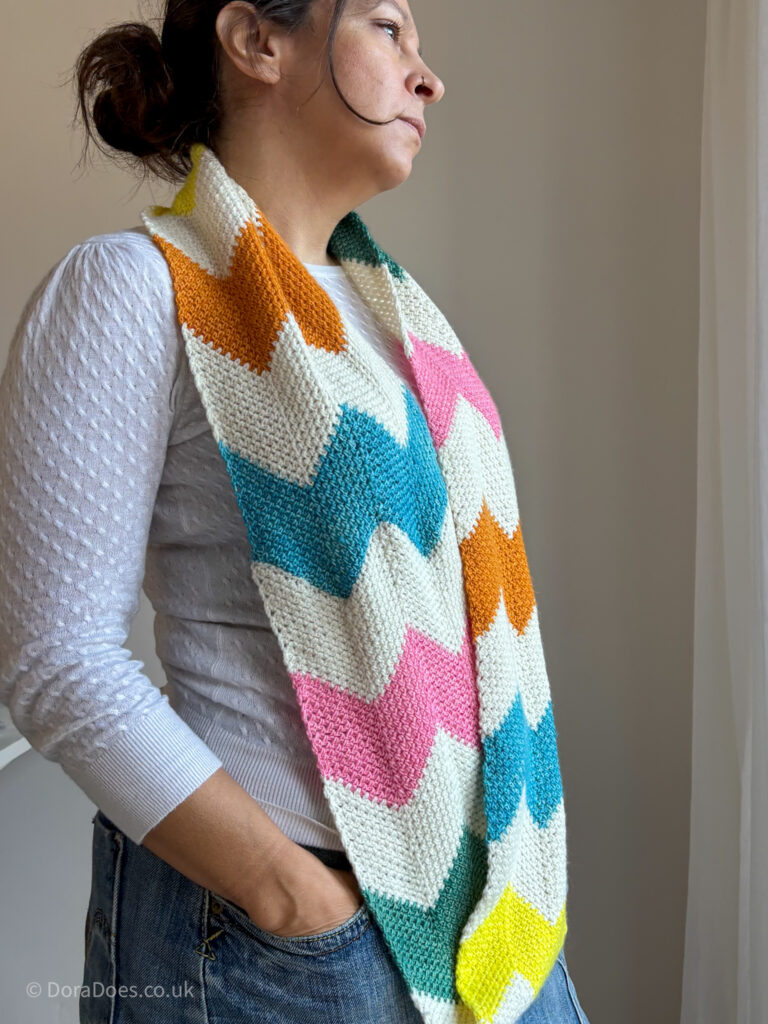 Dora wears a stripy crochet infinity scarf draped long around her shoulders. The chevron stripes mix rainbow colours with undyed natural wool yarn.
