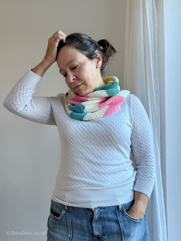 A woman wears a fun, modern crochet scarf with bright pastel zig zag stripes alternating with natural yarn. The scarf is wrapped double and draped over her shoulders. She raises one hand to her head and looks to the floor.