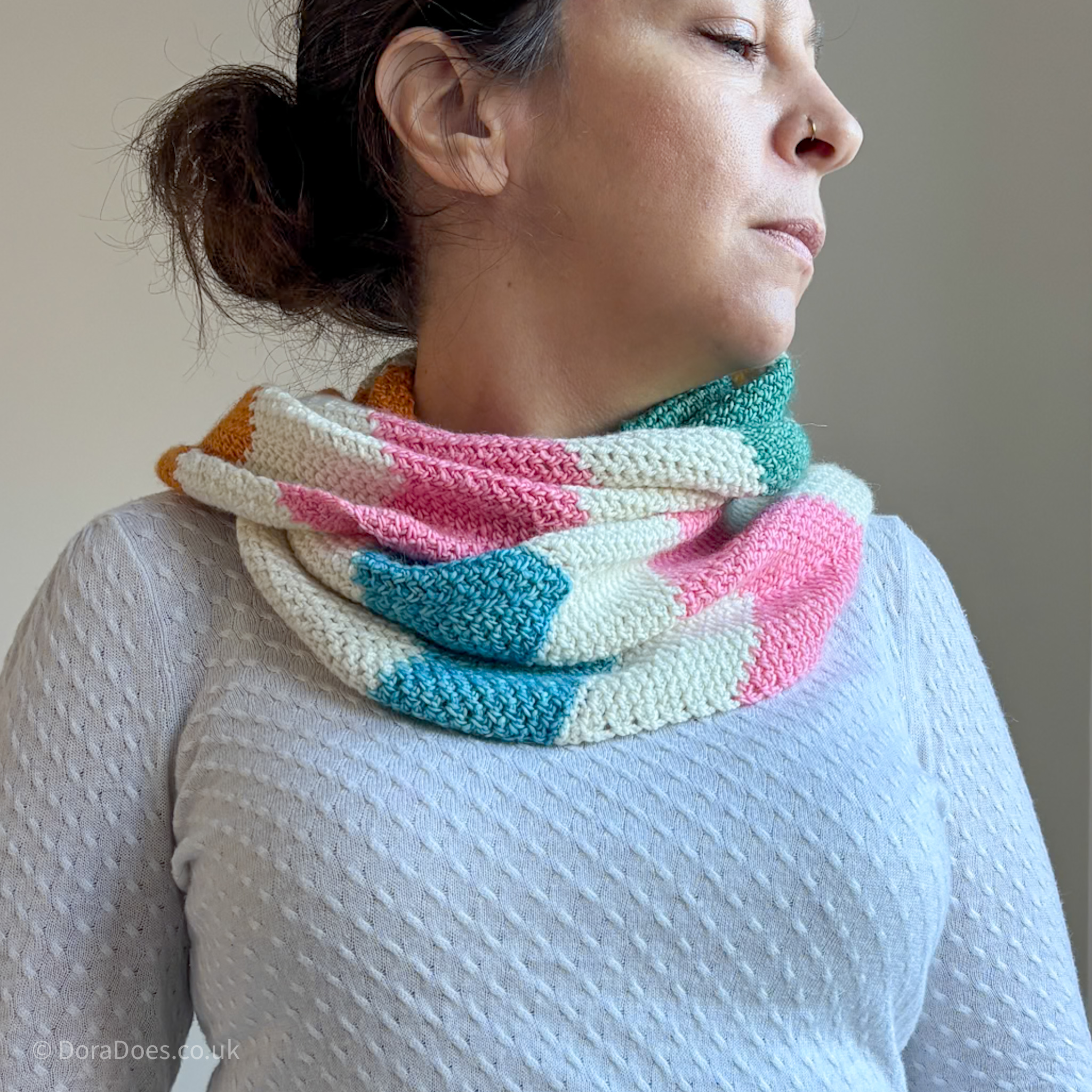 A close up of Dora wearing a 4 ply yarn weight infinity scarf as she looks to one side. The crochet scarf uses moss stitch chevrons to created a chunky zig zag stripe and looks elegant with all its drape and paired with a cream sweater.