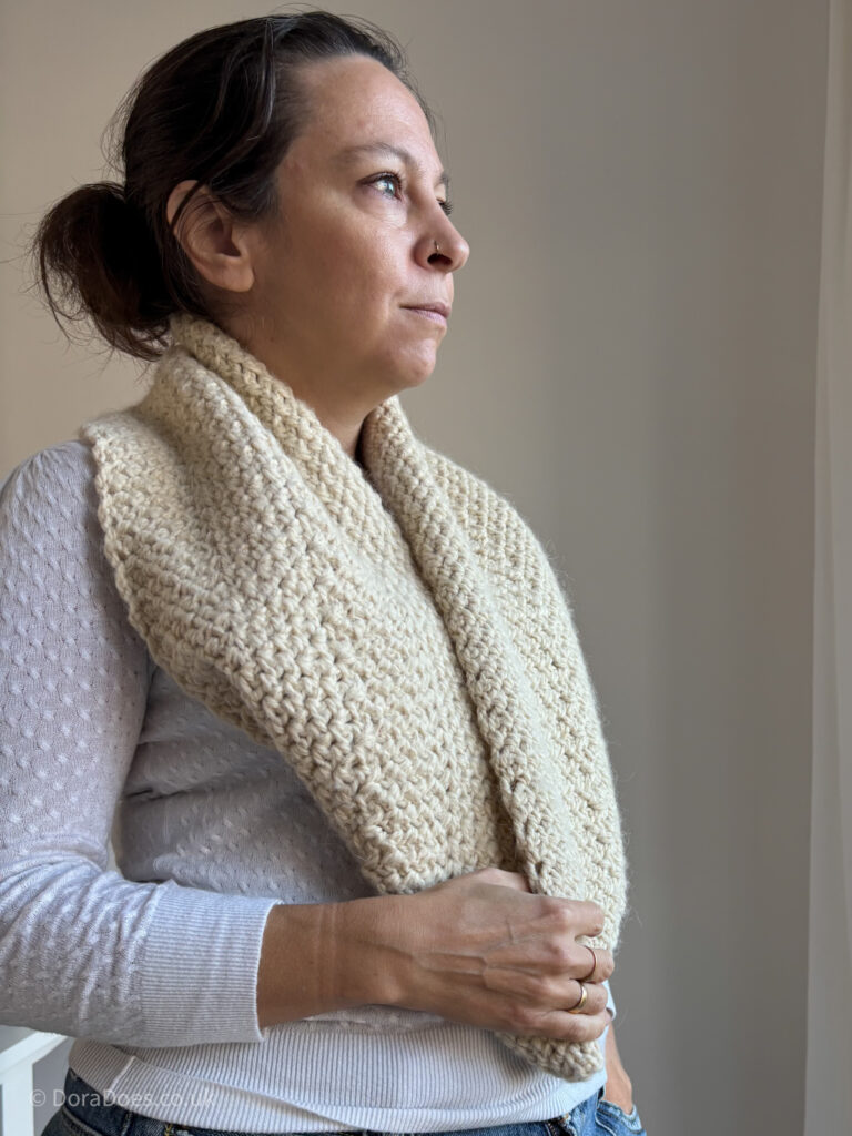 Dora wears a camel colour infinity scarf long, paired with a light cream sweater. The scarf uses a textured crochet stitch with a slightly fluffy yarn making it look warm and inviting