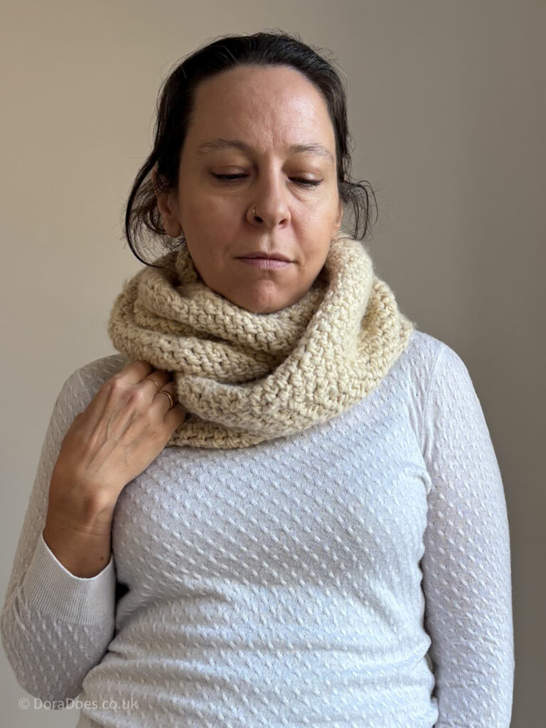 Dora wears an Aran weight crochet infinity scarf wrapped double like a cowl. She holds the bottom of one side of the scarf, emphasising its cozy warmth.