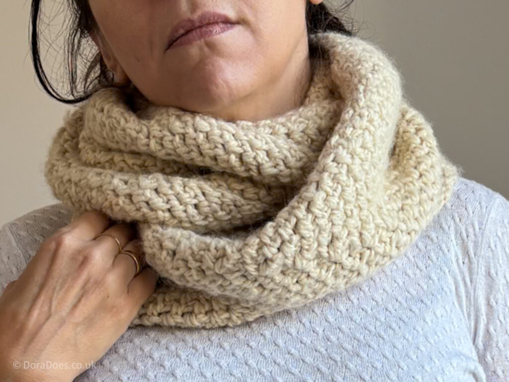 A close up of a crochet infinity scarf work double round a woman’s neck. The natural coloured yarn is crocheted up in a tweed stitch with a subtle chevron.