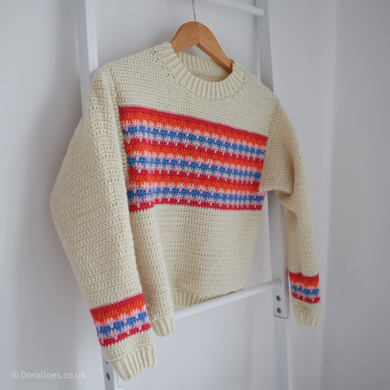 The crochet lark sweater hangs from a blanket latter against a white wall. It's a cream crochet jumper with orange red pink and blue stripes across the chest and matching colour work around the wrists.