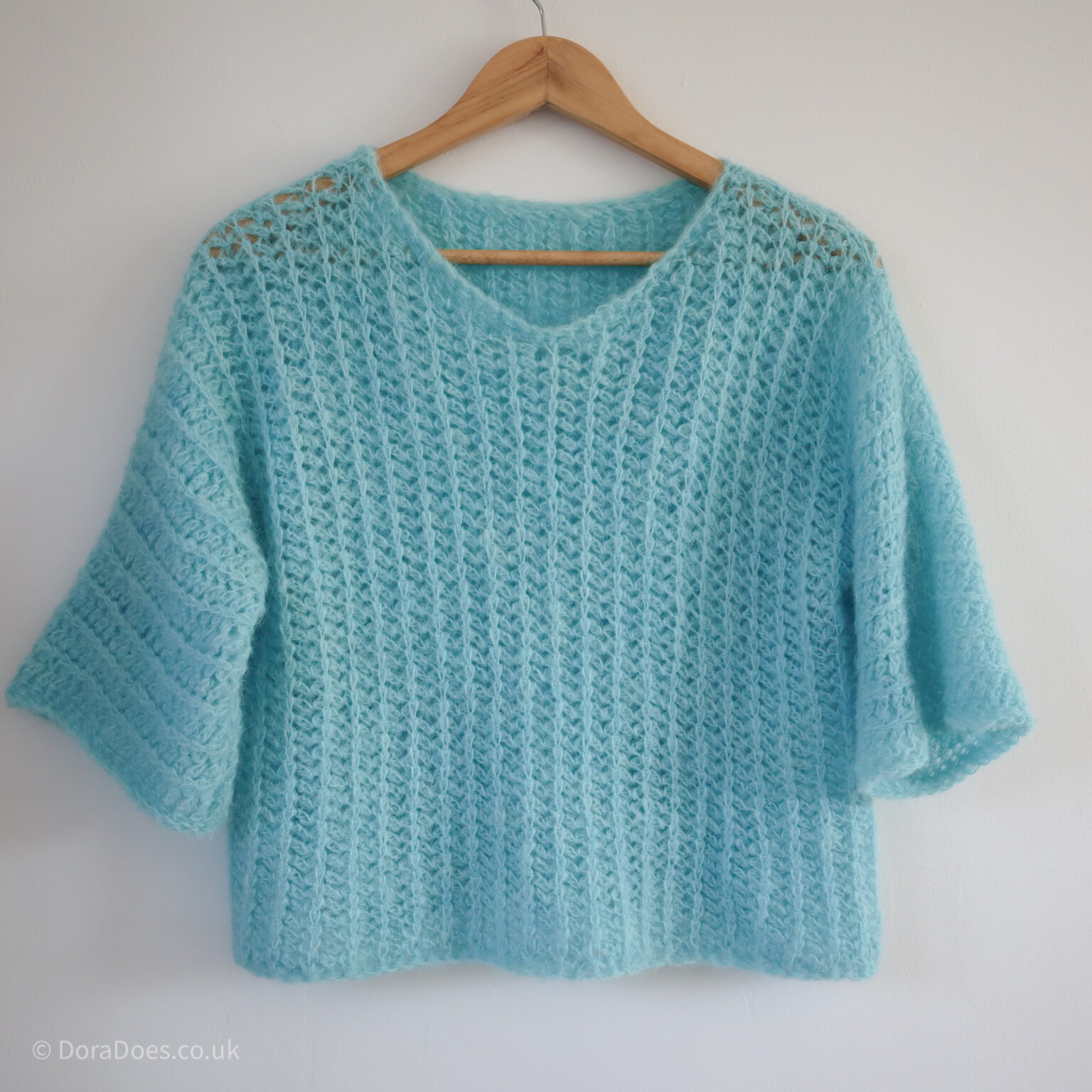 An ice blue fluffy crochet v-neck sweater with relaxed sleeves that are lust less than 3/4 length hangs against a white wall.