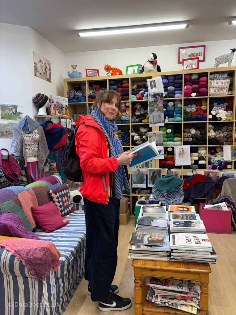 A woman in a bright red jacket stands in the middle of Kathy’s Knits yarn store reading a copy of the Journal of Scottish Yarn.