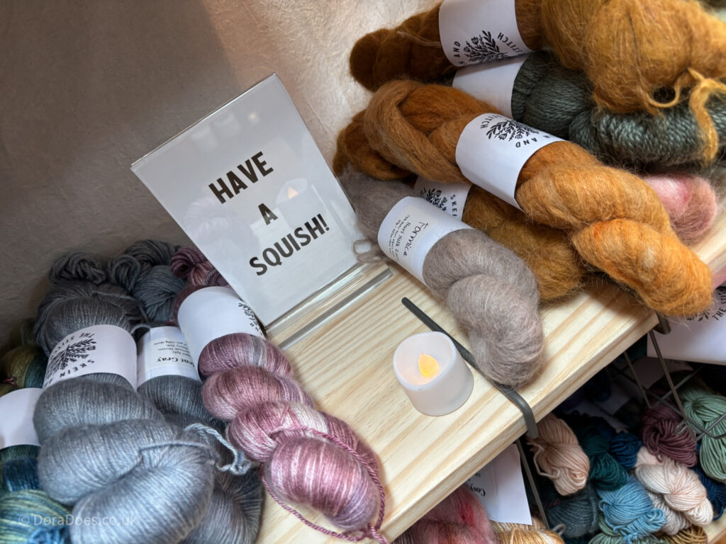 A shelf of hanks of naturally dyed yarn with a sign in between that reads ‘have a squish.’