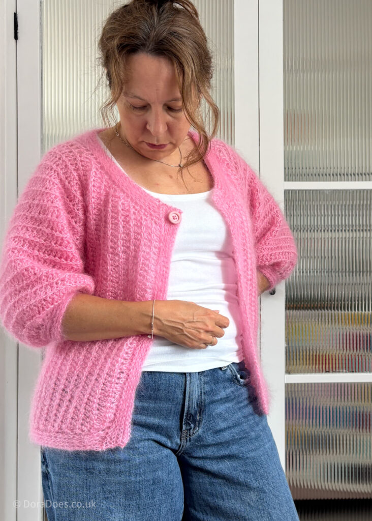 Dora wears a bright pink crochet cardigan with mid-length sleeves and a single pink button, undone, at the neckline. The cardigan drops to har hips and has a ribbed crochet fabric and is worn paired with a white vest top and dark blue wide leg jeans.