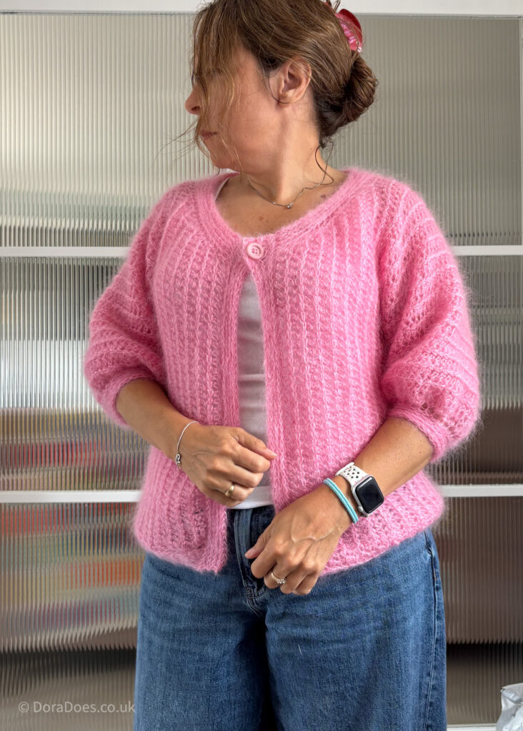 Dora wears the fluff it cardigan paired with jeans as she looks to the side. Seen from the front, the cardigan is fastened with a single pink button and has mid length sleeves. It has a fluffy texture from the mohair yarn and a vertical crochet rib texture in the fabric.