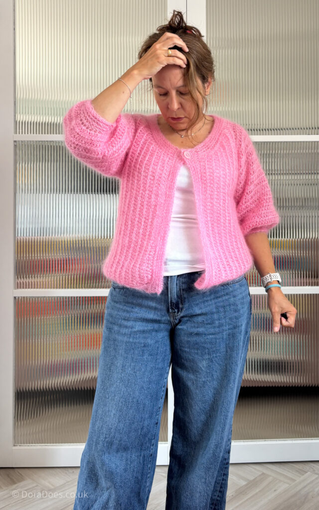 A portrait image of Dora seen from the front wearing a pink fluffy crochet cardigan with a single button at the top paired with a white tank and jeans.