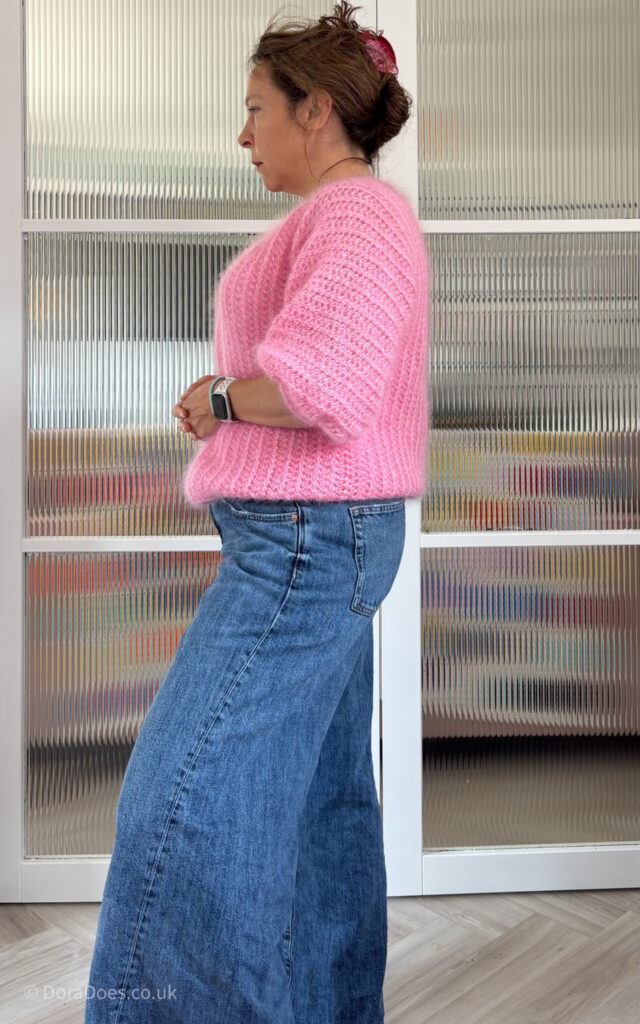 Dora is seen from the side wearing a pink bomber jacket style crochet cardigan made from pink fluffy yarn.