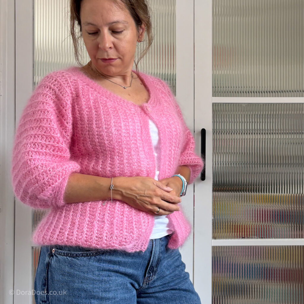 Dora wears the ‘Fluff it’ crochet cardigan, a bright pink fluffy bomber jacket shaped cardigan with mid length sleeves and a ribbed knit-look texture.