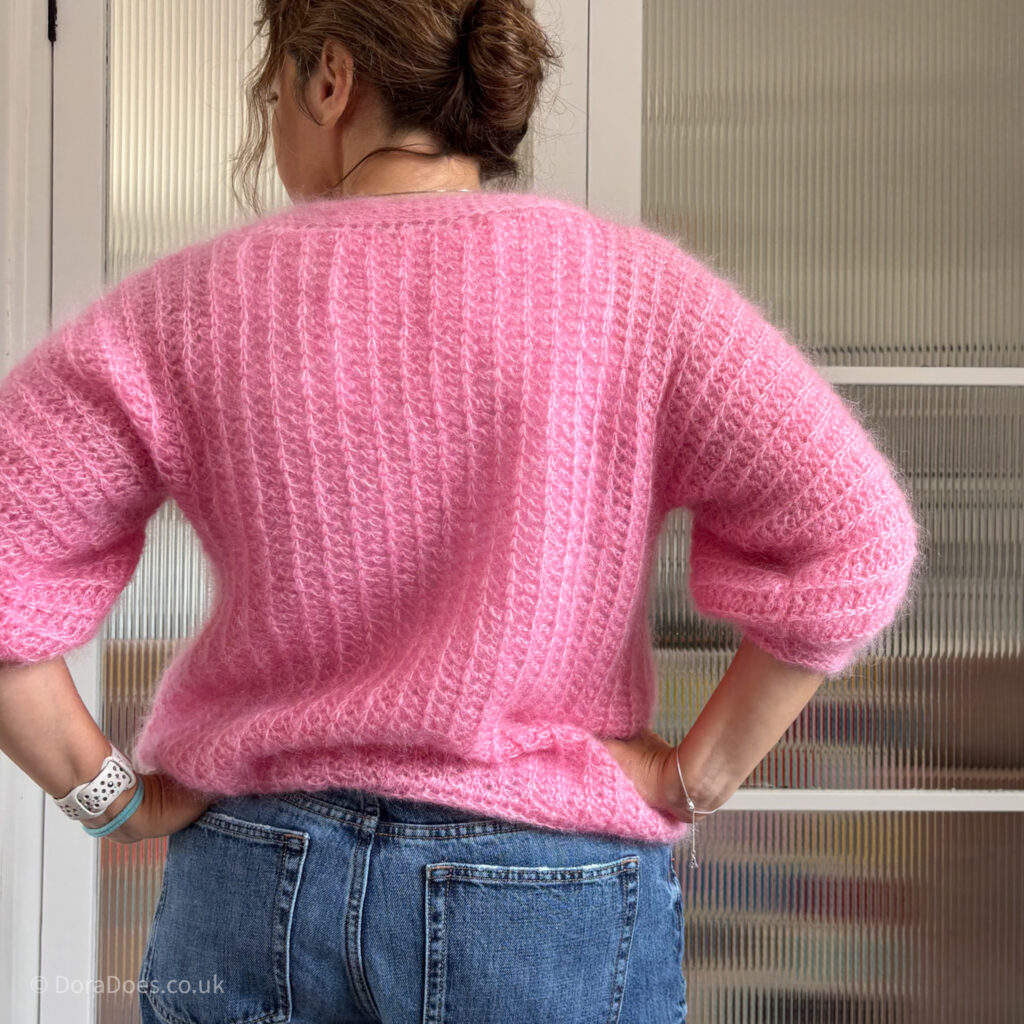 Dora is seen from behind, standing with hands on hips wearing the ‘fluff it’ crochet cardigan; a ribbed knit look crochet jacket made with mohair and silk yarn.
