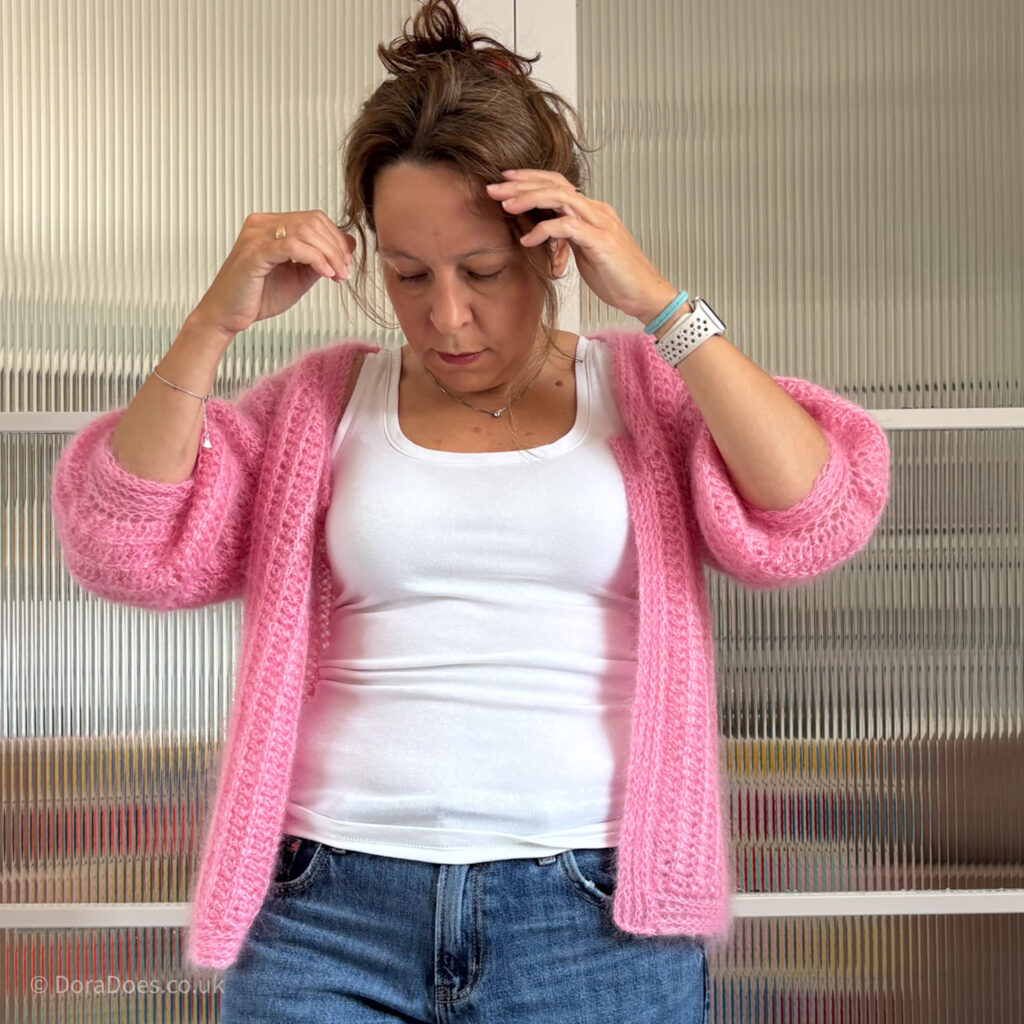 Dora wears a pink fluffy crochet cardigan with a vertical ribbed texture and sleeves that fall just below the elbow.