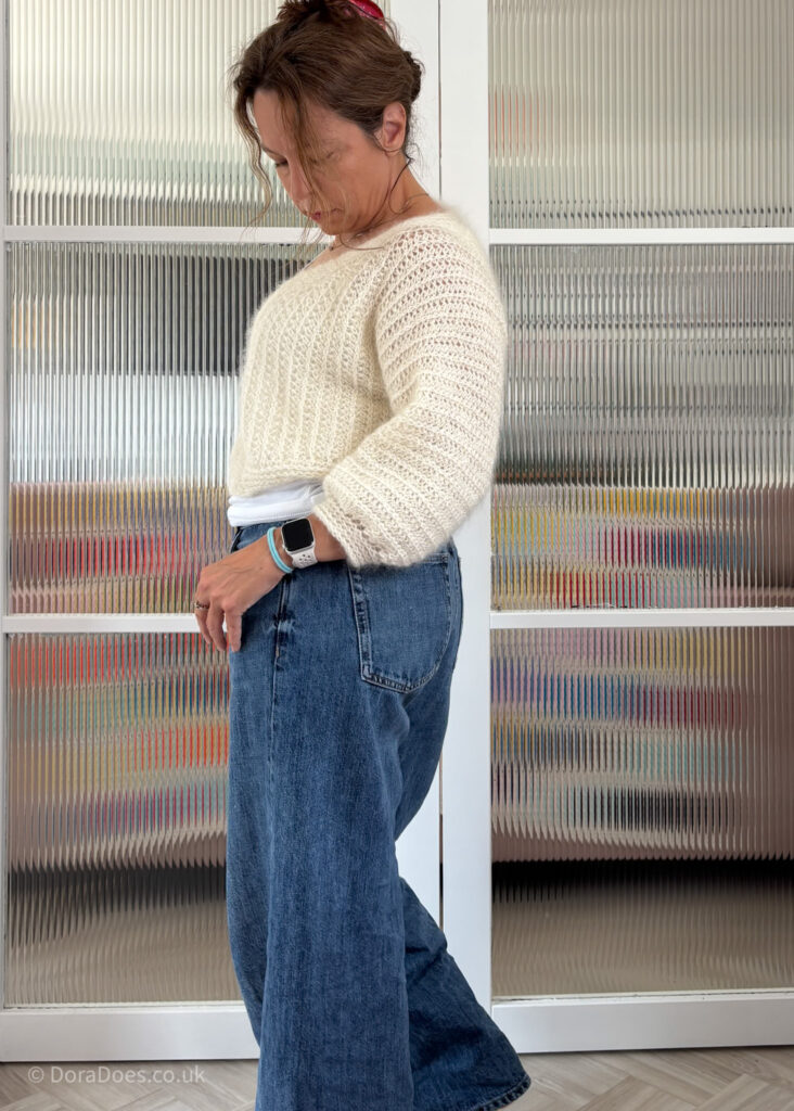 A side view of Dora wearing a cropped fluffy cream cardigan made from two strands of undyed yarn held together to create a vertical rib stitche. The cardigan is paired with wide leg jeans.