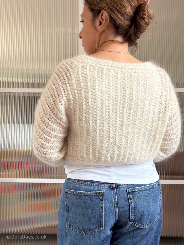 Dora is seen from behind wearing a cream, cropped version of the ‘fluff it’ cardigan; a ribbed knit look crochet cardigan.