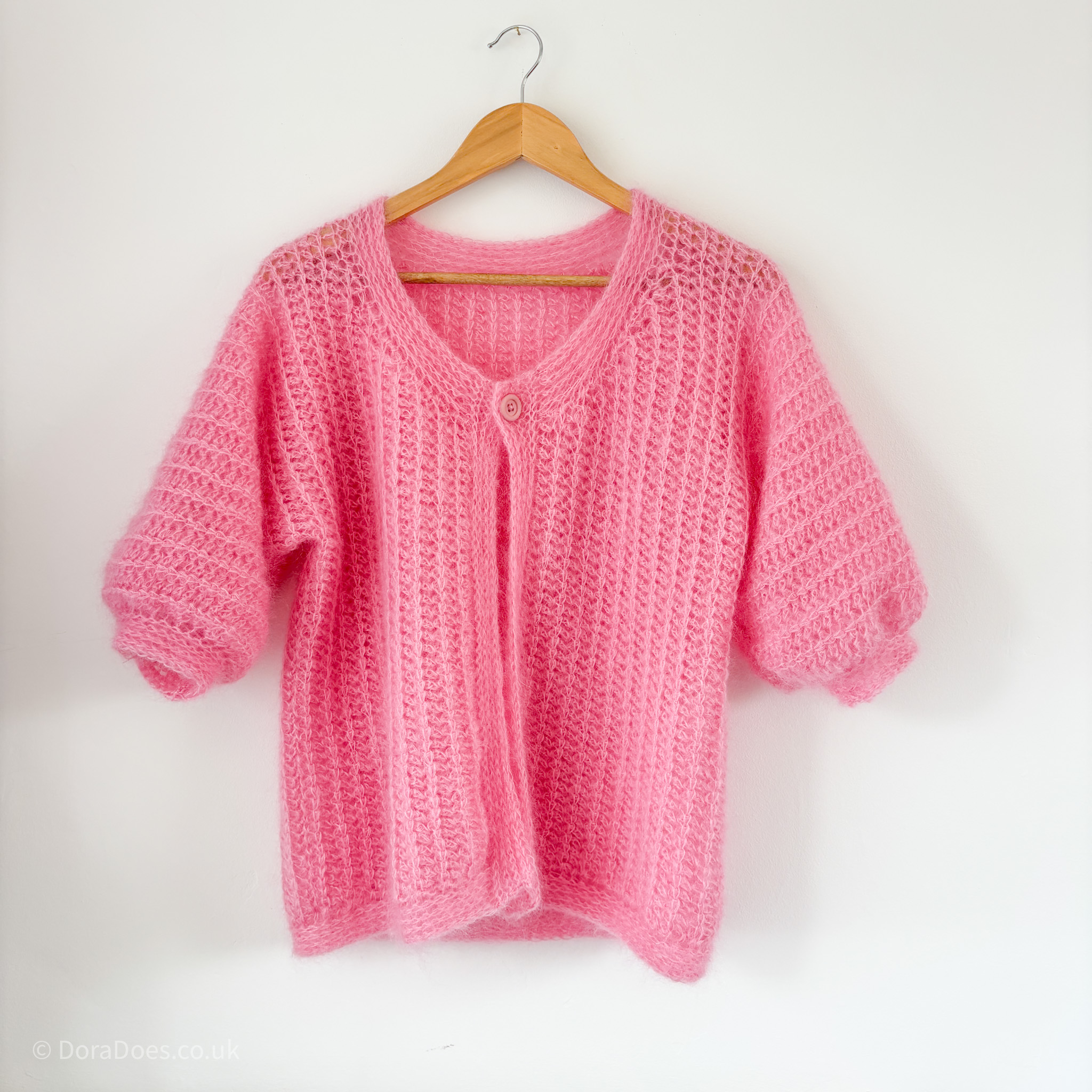 A pink mohair crochet cardigan hangs against a white wall. The cardigan has 2/3 length sleeves with a tightly ribbed collar, button band and hem. The crochet fabric drapes nicely and is made with a crochet rib stitch.