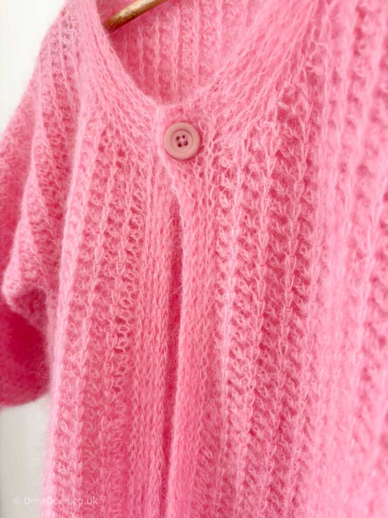 A close up of the neckline and button band of the ‘fluff-it’ crochet cardigan showing the single pink button at the neck opening and the knit look texture of the collar and button band, made tighter than the ribbing in the rest of the crocheted fabric.