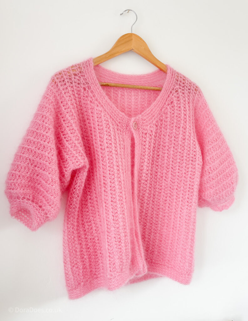An angled view of the ‘fluff it’ cardigan hanging against a white wall. The cardigan is made from mohair silk yarn with a vertical crochet rib stitch. With a single button at the neckline and mid-length sleeves, it has lots of drape and a knit-look trim.