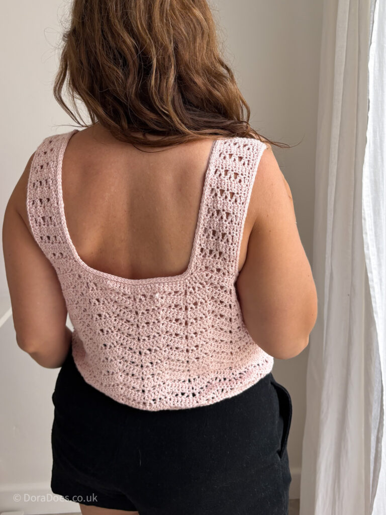 Dora is seen from behind wearing a pink crochet vest top. The top is made with 4ply wool and silk yarn and is made with a crochet wave stitch in lacy patterns.