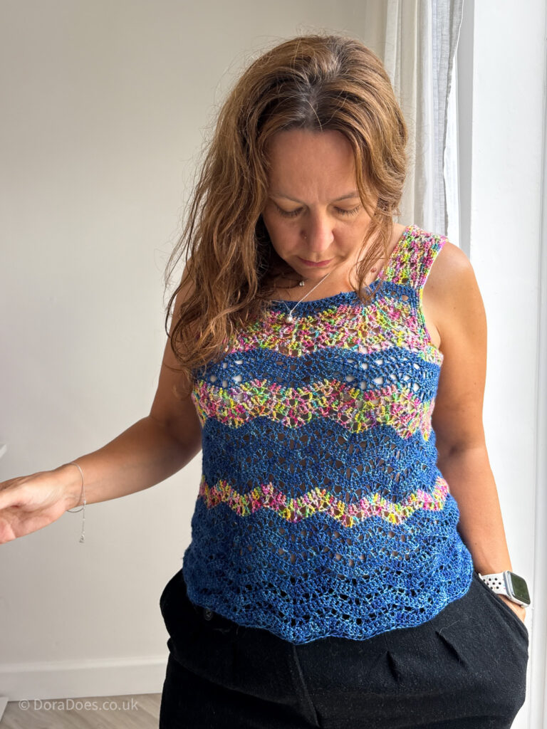 Dora looks down to admire the blue and rainbow crochet tank top she is wearing. The crochet vest has horizontal zig zag stripes in dark blue and variegated rainbow yarn.